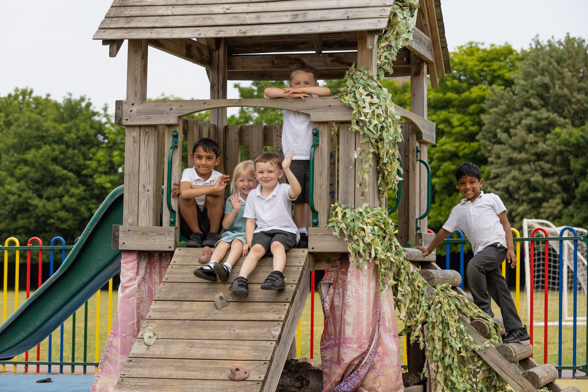 Pupils sitting/leaning on wooden play equipment while smiling and waving at the camera